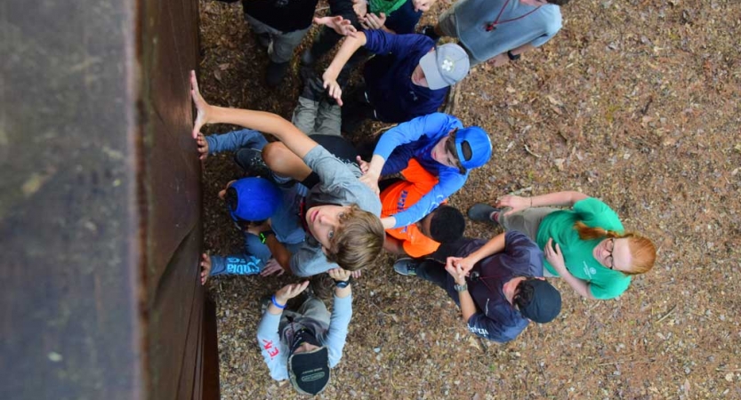 From the top of an obstacle, the camera looks down at a group of students helping another up.